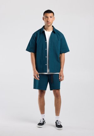 Jonge man die recht vooruit kijkt, gekleed in een turquoise kort mouw overhemd met knopen, bijpassende korte broek, wit T-shirt, witte sokken en zwarte sneakers.