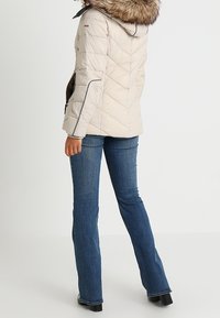 Jasno beżowa kurtka ocieplana z kapturem z sztucznego futra, pikowany wzór i ciemnobrązowe boczne kieszenie. Noszona z dzwonowymi niebieskimi dżinsami.