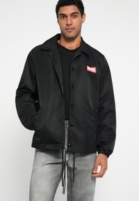 Μαύρο σακάκι προπονητή από νάιλον με κουμπιά με πατητό μηχανισμό, γιακά και κόκκινο λογότυπο στην αριστερή πλευρά του στήθους. Διαθέτει ελαστικά μανίκια και ρίγα με κορδόνι στο κάτω μέρος.