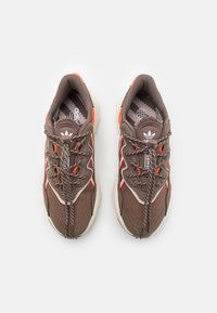 Pár hnědých tenisek Adidas s oranžovými a červenými akcenty, vzorovanou vnitřní stélkou a vzorovanými tkaničkami svázanými volným uzlem.