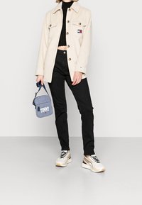 Beżowa polarowa kurtka z guzikami i kieszeniami, czarne dopasowane dżinsy oraz białe trampki z beżowymi akcentami. Niebieska torba z logo "TOMMY".