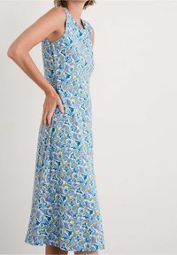 Kobieta w bezrękawowej sukience do kolan w niebiesko-fioletowo-żółty kwiatowy wzór, stojąca bokiem, z jedną ręką na biodrze.