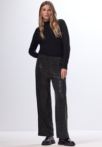 Schwarzer gerippter Rollkragenpullover kombiniert mit schmalen schwarzen Hosen aus Pailletten in High-Waist-Form. Das Model trägt klobige schwarze Schuhe. Schlichter Hintergrund.