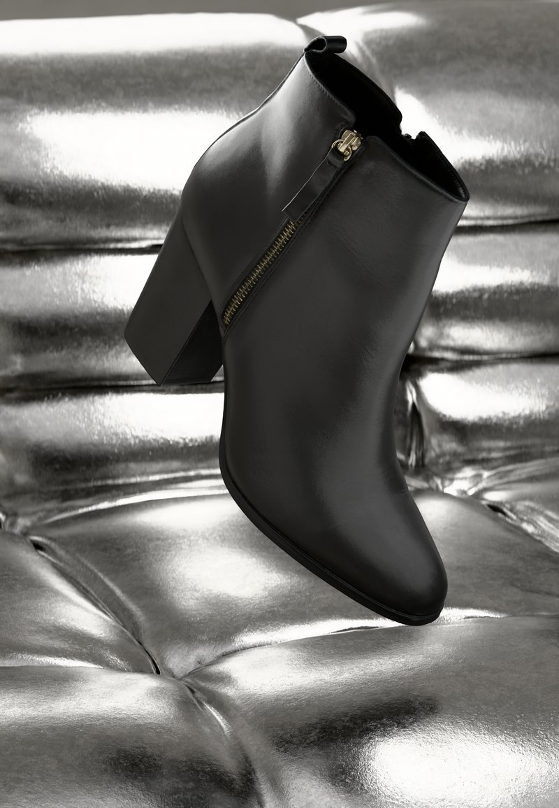 Schwarze Leder-Stiefelette mit spitzer Zehenform, Blockabsatz und goldenen Reißverschlussdetails. Vor einem silbernen strukturierten Hintergrund. 