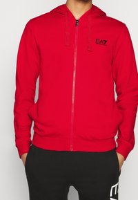 Sudadera con capucha roja con cremallera, mangas largas, capucha con cordón ajustable y un pequeño logo negro en el pecho izquierdo. Fabricada con tela suave.