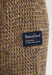 Close-up van een geelbruin gebreide stof met een marineblauw label met de tekst "Petrol Ind, Denim Driven, Ref. 5026 Sinds '89 U.S.A."