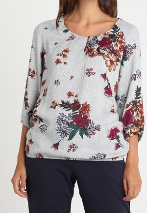 Blouse florale en tissu gris clair avec un col rond, des manches trois-quarts et des motifs de fleurs colorées dans des teintes bourgogne et jaune.