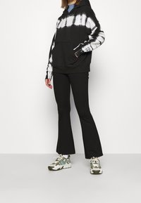 Sudadera negra con efecto tie-dye y patrón blanco, con bolsillo frontal. Combinada con pantalones de campana negros y zapatillas con patrón blanco y verde.