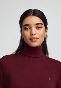 Junge Frau mit dunklem Haar, die goldene Creolenohrringe und einen bordeauxfarbenen Rollkragenpullover mit kleinem gesticktem Logo auf der Brust trägt.