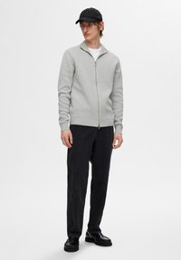 Un pull zippé gris clair en tricot doux, associé à un jean noir à jambes droites et des chaussures noires, sur fond blanc uni.
