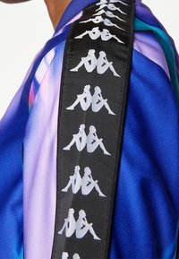 Cintura nera con il logo Kappa bianco ripetuto che presenta due figure sedute schiena contro schiena su una maglietta sportiva con sfumature blu e viola.