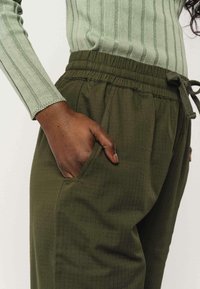 Olijfgroene, gestructureerde joggingbroek met een elastische tailleband en trekkoord. Voorzien van zijzakken en een relaxte pasvorm, gecombineerd met een geribbelde, mintgroene top.