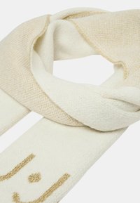 Knitted scarf in cream and light beige with a textured pattern. Features gold threading and a playful design at one end.
