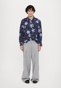 Camisa de manga larga de un azul marino oscuro con un patrón floral azul; combinada con pantalones grises claros con pinzas y zapatos negros. Accesorio de bolso de hombro marrón.
