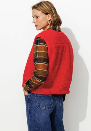 Veste en polaire rouge avec une surface texturée, portée sur une chemise à carreaux. Le mannequin a des cheveux mi-longs et est associé à un pantalon en denim.