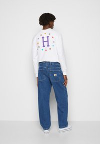 Mies, joka ihailee lyhyitä dreadlokkeja, on pukeutunut valkoiseen pitkähiharaiseen paitaan, jossa on purppura 'H' ja monivärisiä tähtiä selässä, sinisiin farkkuihin ja valkoisiin lenkkareihin.