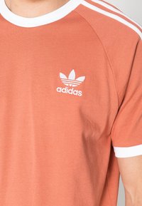 Triko s krátkým rukávem a kulatým výstřihem v barvě lososu, vyrobené z bavlny, s bílými akcenty a logem Adidas na hrudi.