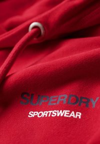 Felpa rossa con una texture morbida. Presenta occhielli in metallo argentato per il cordino e la stampa di "SUPERDRY" e "SPORTSWEAR" in colori a contrasto.