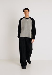 Langärmliger, gerippter grauer und schwarzer Pullover mit Rundhalsausschnitt, kombiniert mit lockeren schwarzen Hosen und klobigen schwarzen Schuhen.