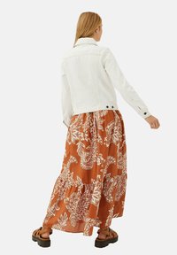 Witte denimjack over een oranje maxi-jurk met witte bloempatronen. De jurk heeft een gelaagd ontwerp en is gecombineerd met bruine sandalen.