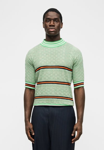 Muž nosiaci svetlozelený texturovaný sveter s krátkym rukávom so čiernymi, oranžovými a zelenými pruhovanými pásmi, v kombinácii s tmavými nohavicami s jemnými pruhmi.