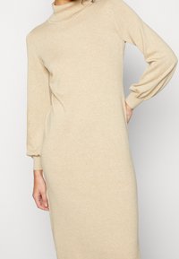 Beżowa dzianinowa sukienka z wysokim kołnierzem, długimi puff rękawami i dopasowanym krojem, o miękkiej fakturze i minimalistycznych akcentach wzorniczych.