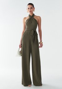 Combinaison olive avec un col halter, détail de taille à nouer, design à jambes larges et tissu texturé. Les accessoires comprennent un sac à main blanc.