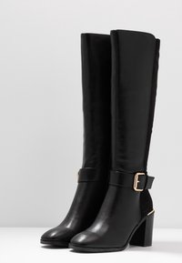 Black knee-high boots made from leather and suede, featuring a chunky heel, wide strap with a gold buckle, and a smooth texture.