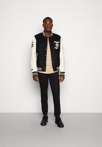 Black and cream varsity jacket with green embroidery, striped cuffs, and black cargo pants, paired with black sneakers. Model stands upright.