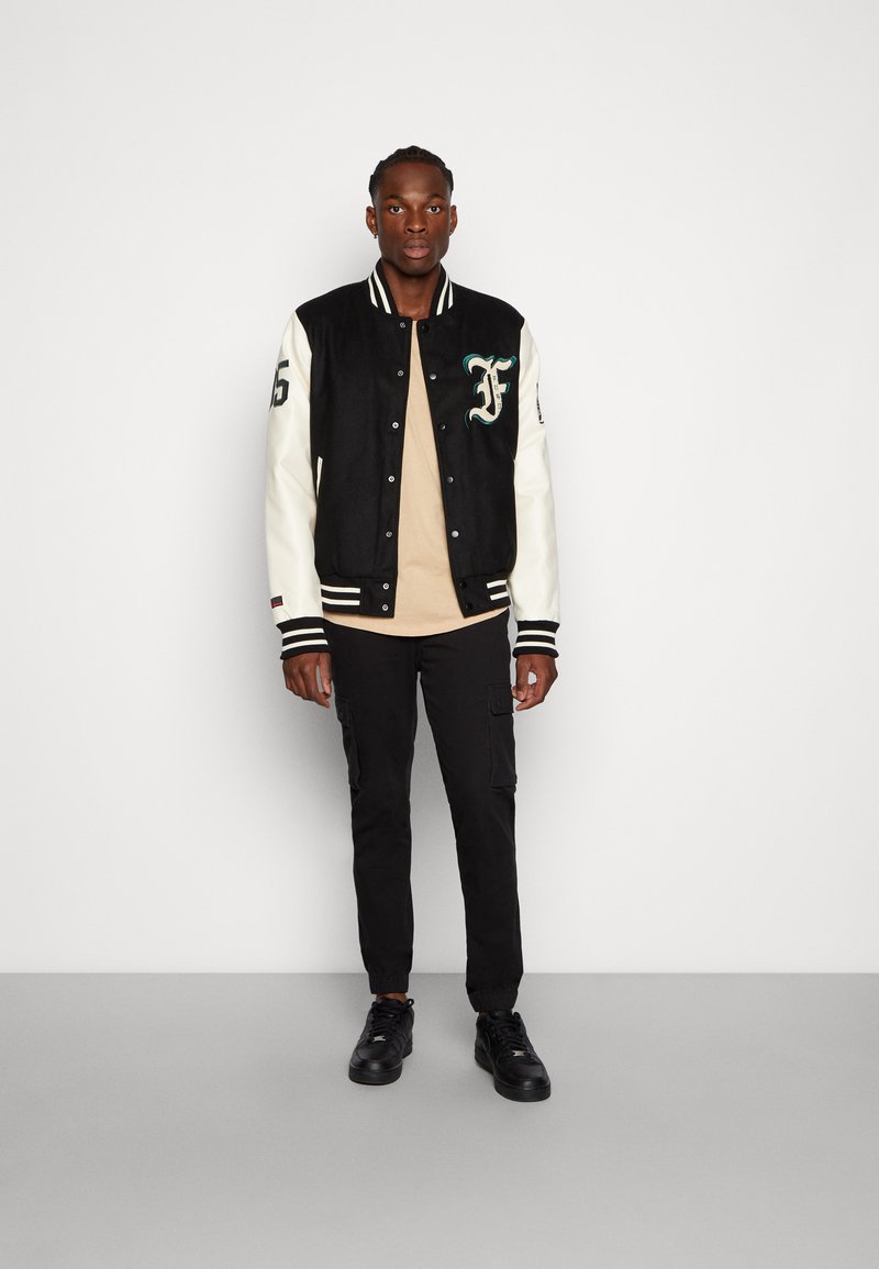 Black and cream varsity jacket with green embroidery, striped cuffs, and black cargo pants, paired with black sneakers. Model stands upright.