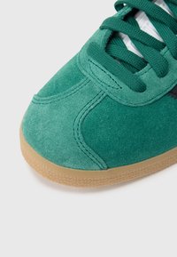 Sapatilha verde de camurça com superfície texturizada, sola de borracha cor de goma, atacadores proeminentes e detalhes subtis de costura.