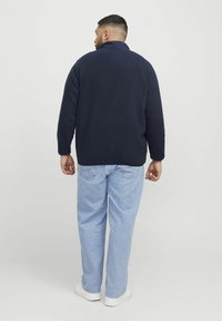 Marine fleecejakke med høj krave, raglanærmer og en struktureret overflade, parret med lyseblå denimjeans og hvide sko.