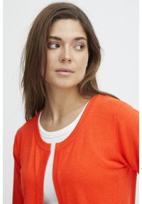 Cardigan orange avec un col rond, des bordures côtelées et une fermeture à boutons. Porté par-dessus un haut blanc. Texture douce, design léger.