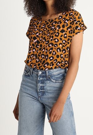 Nainen, joka pukeutunut lyhythihaiseen, ruskeaan leopardi-printtipaitaan, joka on sullottu vaaleansinisiin, korkeavyötäröisiin haalistuneisiin farkkuihin, seisoo toinen käsi sivulla.