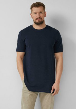 Homme avec une barbe portant une chemise à manches courtes bleu marine et un pantalon beige, debout devant un fond gris clair uni.