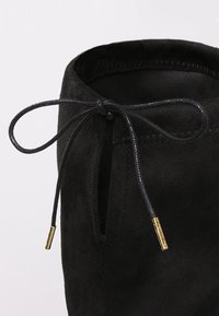 Tige de chaussure en daim noir avec un lacet noué en forme de nœud, doté d'œillets métalliques dorés sur un fond clair et neutre.