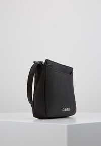 Černá syntetická kabelka se strukturovaným designem, s vrchním zipem, nastavitelným popruhem a stříbrným logem na přední straně.