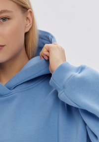 Felpa azzurra chiara con un tessuto morbido e texturizzato, dotata di un grande cappuccio e polsini a costine. Il materiale appare accogliente e rilassato.