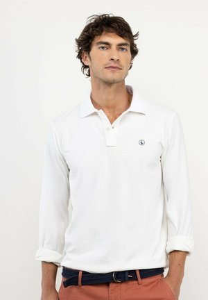 Hombre con cabello corto y rizado que lleva una camisa tipo polo blanca de manga larga y pantalones de color óxido con un cinturón oscuro, de pie contra un fondo liso.