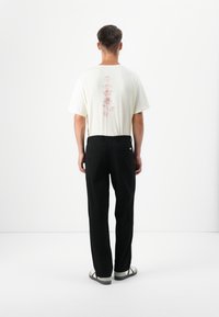 A man stands facing away, wearing a cream t-shirt with red floral graphics, black pants, and sandals, against a plain white background.