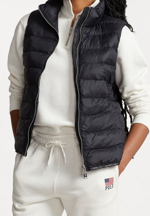 Personne portant un sweat-shirt blanc à fermeture éclair quart, un pantalon de survêtement avec un gilet matelassé noir par-dessus, les mains dans les poches, logo Polo avec drapeau sur le pantalon.