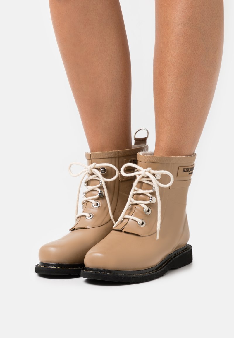 Botines tobilleros de color marrón claro con acabado brillante, cordones blancos, ojales plateados y una suela negra resistente. Presenta un forro suave texturizado y una pestaña de agarre.