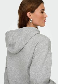 Femme en sweatshirt à capuche gris avec des boucles d'oreilles en forme de cercle en argent, présentée en vue de profil sur un fond clair uni.