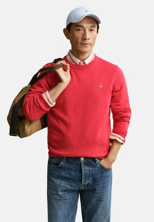 Homme portant un pull rouge par-dessus une chemise rayée, un jean bleu et une casquette blanche, tenant la sangle d'un sac de voyage marron sur l'épaule.