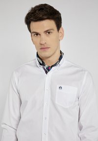 Biała koszula z guzikami z małą kieszenią i granatowym logo na klatce piersiowej. Kołnierz noszony jest z wzorzystym szalikiem na szyi.