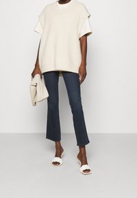 Beige stickad oversized väst med V-ringning, lagd över en vit t-shirt. Mörkblå utsvängda jeans och vita flätade sandaletter.