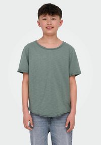 Koszulka z krótkim rękawem, luźna, w jasnym zielonym kolorze z bawełny, o szerokim dekolcie i podwiniętych rękawach, zestawiona z jasnoniebieskimi dżinsami.