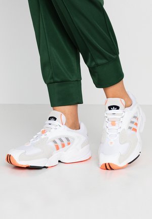 Personne portant des baskets Adidas blanches et orange avec un logo noir, associées à un pantalon vert court sur un sol gris clair.