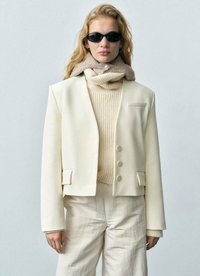 En krämfärgad blazer med slag, tre knappar och fickor, lagd över en beige ribbad tröja. Modellen bär svarta solglasögon.