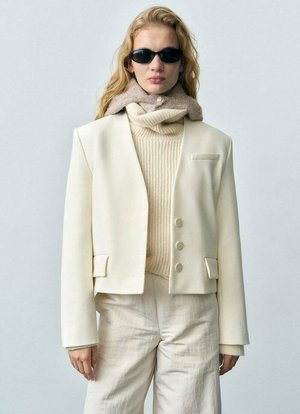 Cream blazer with notched lapels, three buttons, and pockets, layered over a beige ribbed sweater. Model wears black sunglasses.